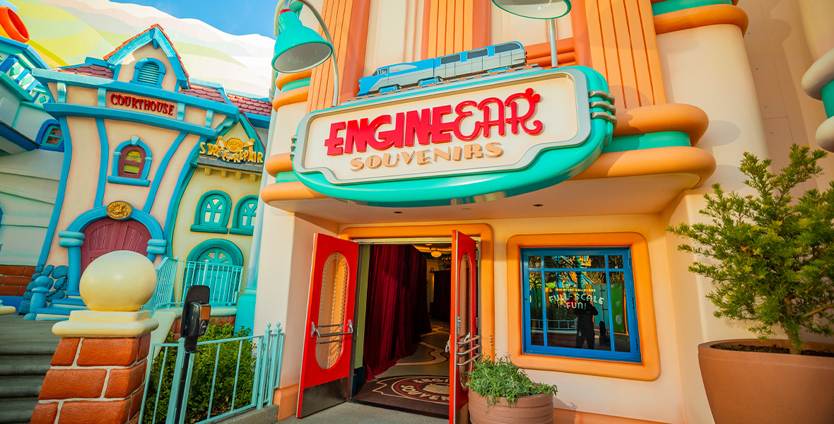 An image of the EngineEar Souvenirs shop at Mickey’s Toontown at Disneyland Park. Its colorful sign above the door has the shop’s name in red script, with two turquoise lamps on top, at either side. On top of the sign itself is a small representation of a train, in blue and gray. Red doors are open to the shop. Several planters are seen to the right, and other Toontown buildings can be seen to the left.