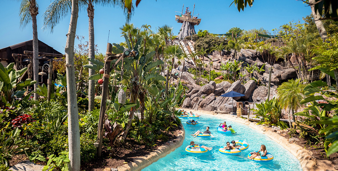 An image of the lazy river from Disney Typhoon Lagoon Water Park at Walt Disney World Resort. Foliage and palm trees line the lazy river, in which guests are sitting in inner tubes floating in the water. The sun is shining. Rock formations can be seen in the background.