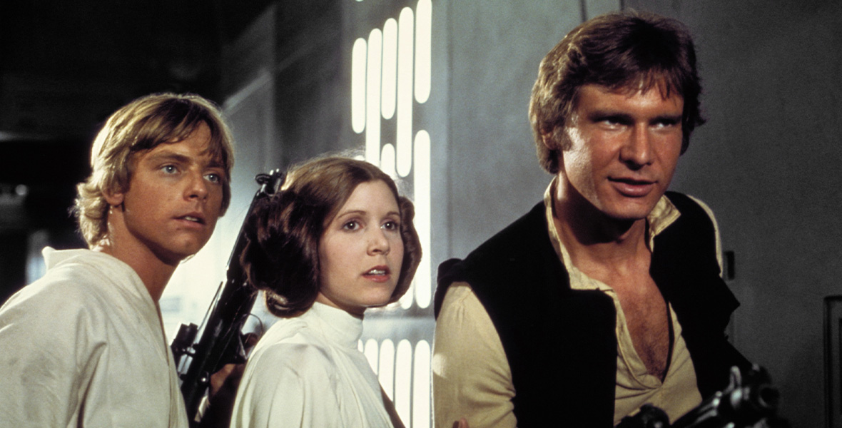 In Star Wars: A New Hope, Luke, Leia, and Han all stand together in front of a metallic wall. Han is positioned slightly in front of the other two and braces his blaster, while Leia stands in the middle and Luke at the left. They look carefully at something ahead.