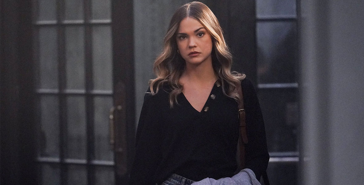 In a scene from an episode of Good Trouble, actor Maia Mitchell stands and holds a blue coat in her left arm. She wears blue denim jeans and a black blouse.