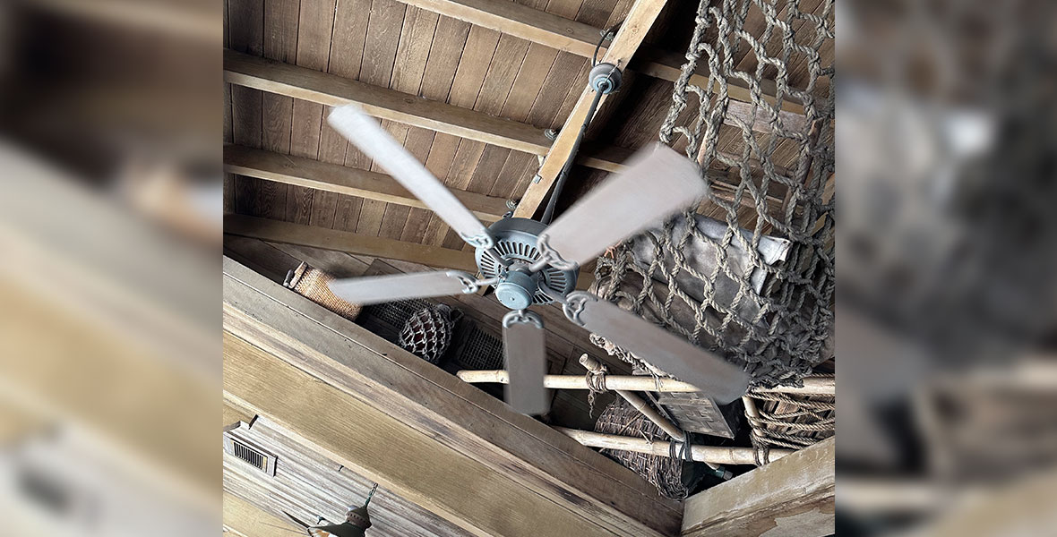 A brown colored ceiling fan hangs from a wood beamed ceiling. Behind the fan is a large net hanging from the ceiling full of older looking cases and artifacts. Other ceiling mounted light fixtures can be seen farther down in the back of the photo.