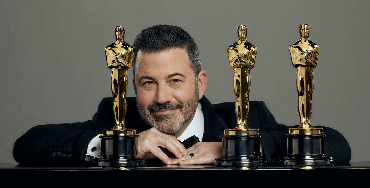 Host and comedian Jimmy Kimmel poses with three Oscars statues and leans on a table.