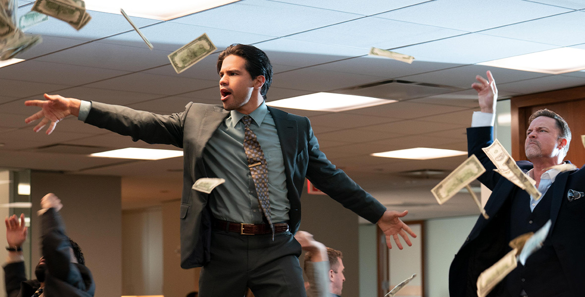 In a scene from an episode of Up Here, actor Carlos Valdes dances on top of a desk inside an office as green dollar bills are thrown around him by actor Scott Porter.