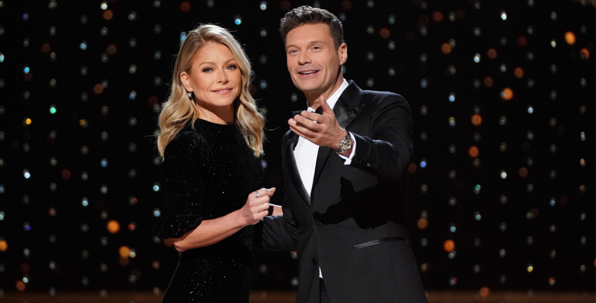 Kelly Ripa and Ryan Seacrest stand together in front of a sparkling black backdrop. Kelly wears a sleek black dress while Seacrest sports a suit and bow tie. Seacrest extends his left hand out as they look slightly off-camera.