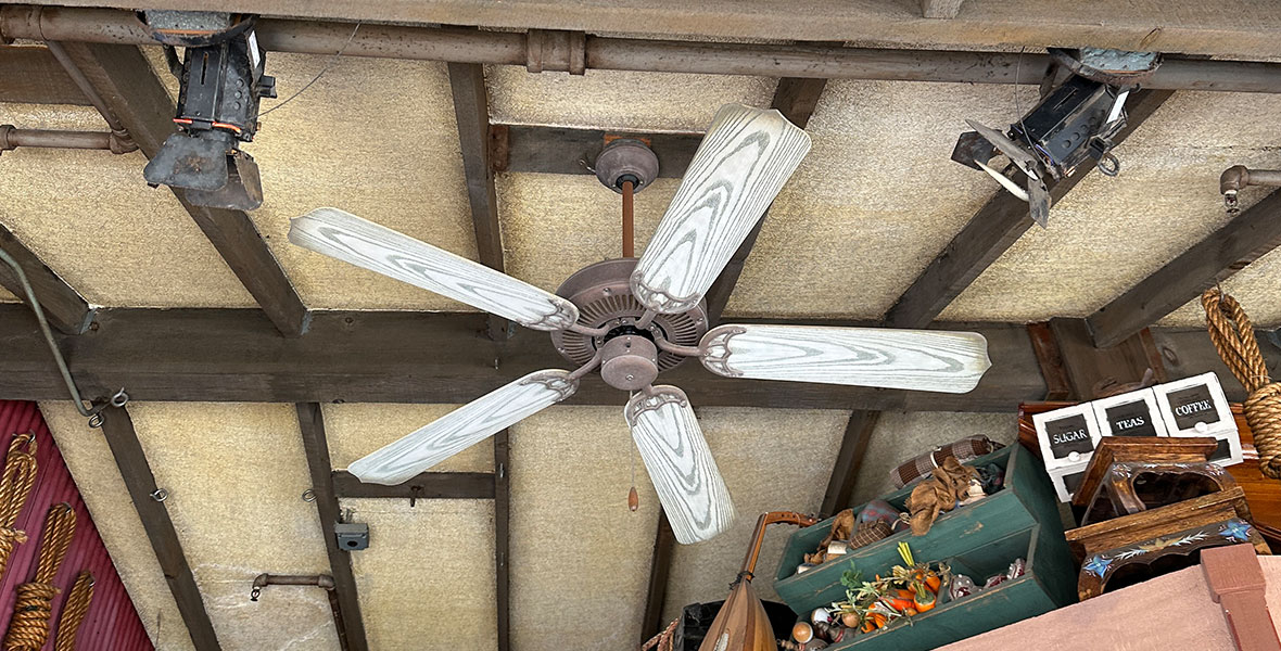 A wooden ceiling fan with blades that appear white-washed from sun exposure hangs from a ceiling that looks like it is made of corkboard with wooden beams. Tons of trinkets and artifacts sit on high shelves close to the ceiling all around the fan. Barely visible in the bottom right corner are Spider Bot toys from Avengers Campus in Disney California Adventure.