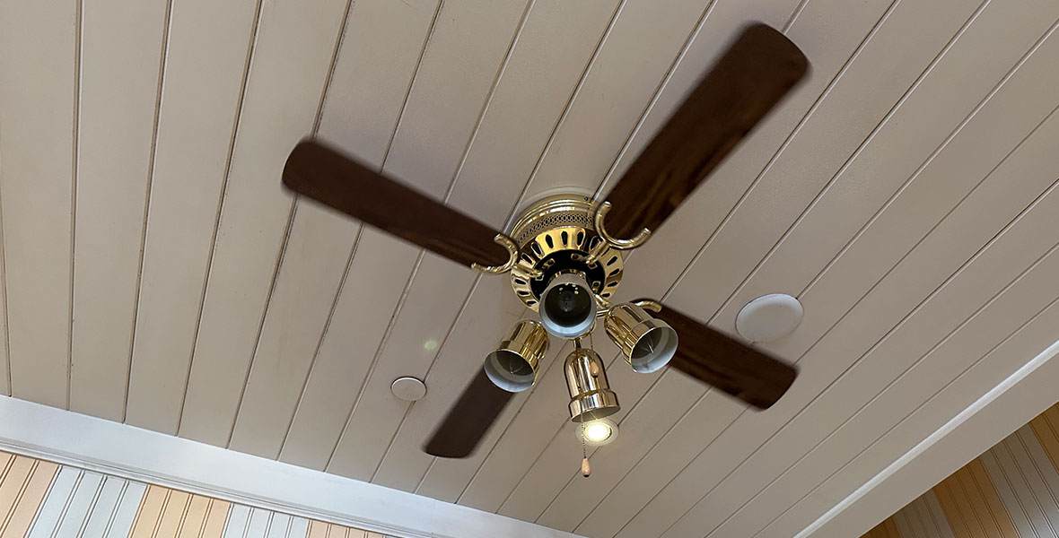 A gold ceiling fan with dark wooden blades is mounted to a wooden slatted ceiling that is painted white. The wall behind the fan is also made of wooden slats that are painted in alternating white and cream colors.