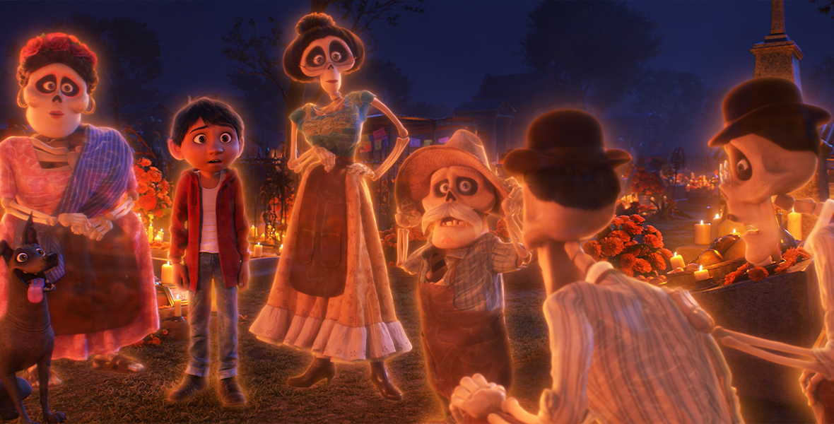 In a graveyard, the young Miguel stands with the five living skeletons of his family members in the animated film Coco. Since they’re all spirits, they’re surrounded by an orange glow. Behind them are several flowers and lit candles as people celebrate Day of the Dead.