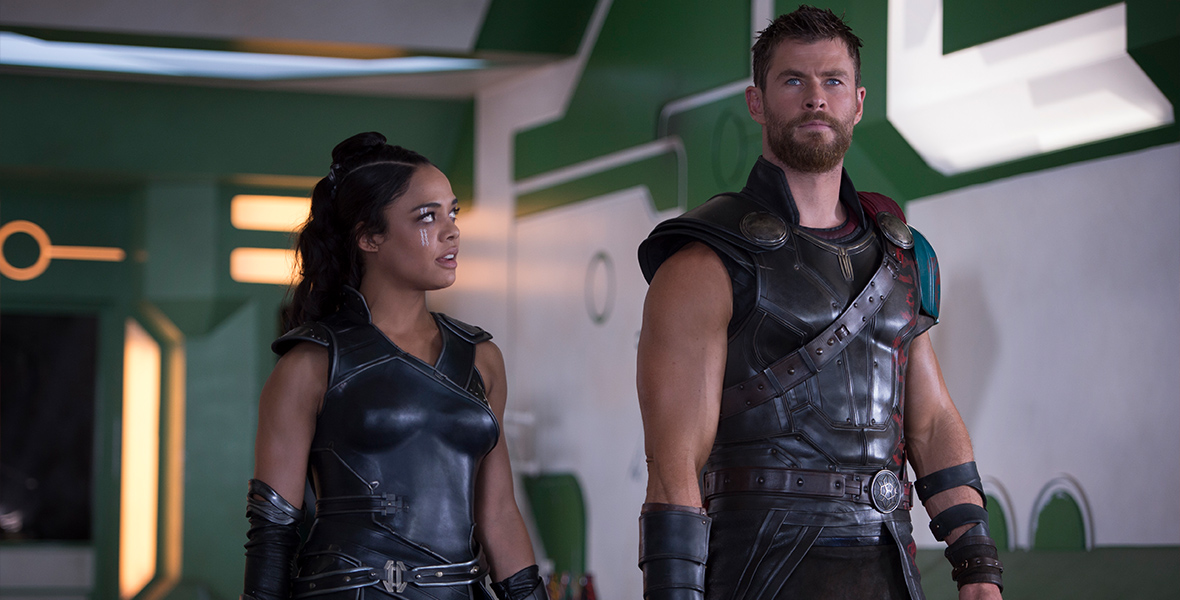 Thor: Ragnarok, Thor and Valkyrie stand beside each other in the Grandmaster’s palace, wearing their warrior armor. Thor’s hair is cut short and he stares into the distance, with Valkyrie looking up at him.