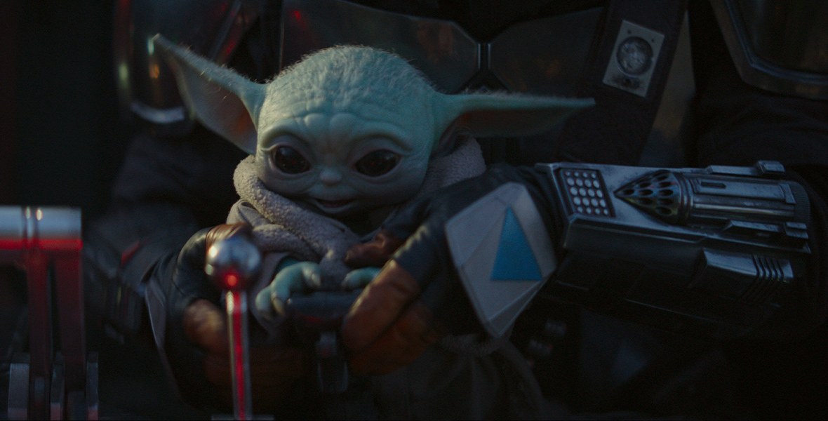 In the series The Mandalorian, a green, Yoda-like infant is held by presumably Din Djarin. Grogu’s large, pointed ears tilt slightly upward and his dark eyes are wide. He smiles.