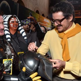 Pedro Pascal, wearing brown glasses, khaki pants, a yellow blouse and a mustard sweater draped over his shoulders, autographs a Mandalorian helmet. Fans who are dressed up in Star Wars cosplay surround Pascal and take photos of the actor.