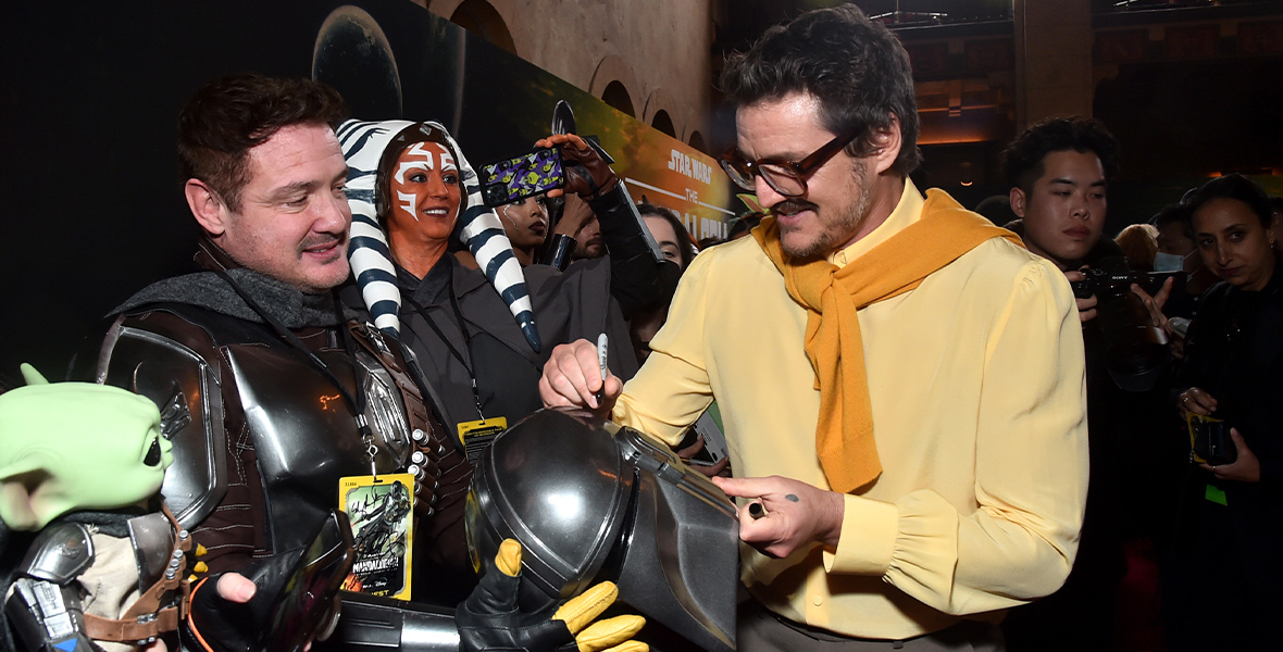 Pedro Pascal, wearing brown glasses, khaki pants, a yellow blouse and a mustard sweater draped over his shoulders, autographs a Mandalorian helmet. Fans who are dressed up in Star Wars cosplay surround Pascal and take photos of the actor.