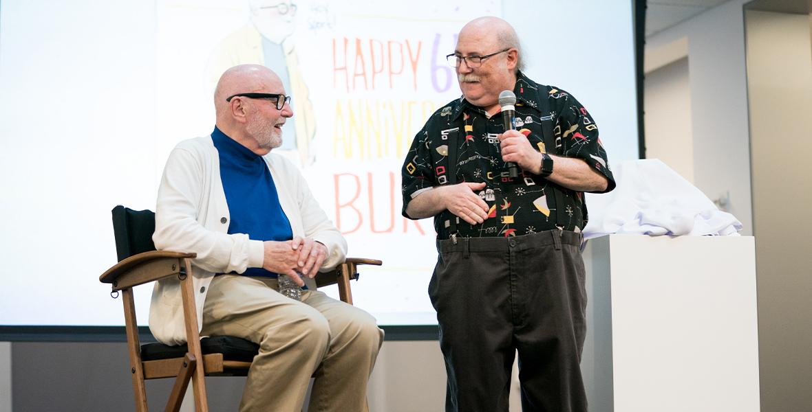 (From left to right) Burny Mattinson sits in a chair on a stage, wearing a bright blue shirt underneath a white cardigan and khaki pants. He wears dark-rimmed glasses and is turned toward animator Eric Goldberg, who has a microphone in hand and is wearing a patterned button-up shirt, dark pants, and eyeglasses. The two men are smiling at one another. A display in the back reads “Happy 65th Burny.”
