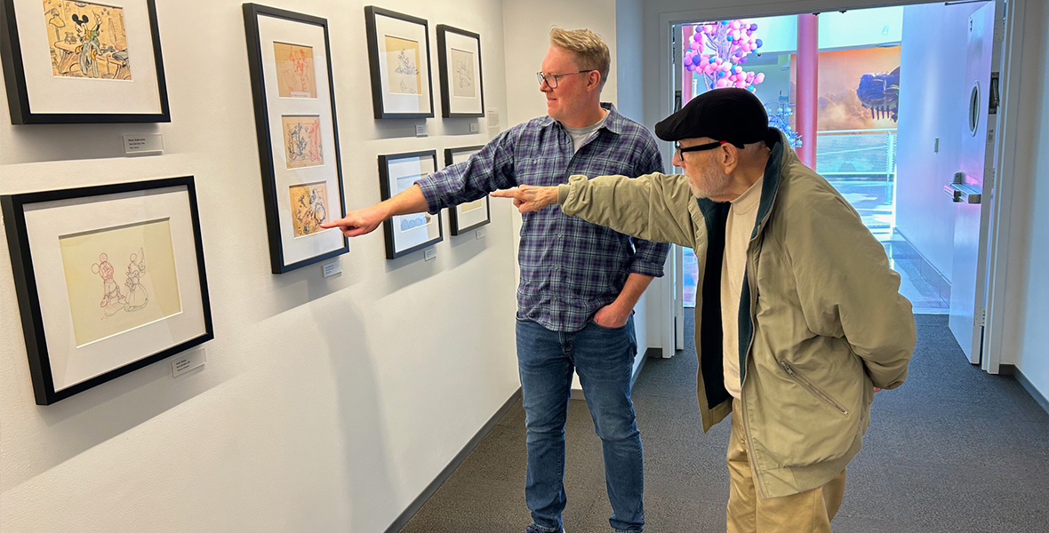 (From left to right) Don Hall points at a detail in a Disney Animation artwork among many framed on the wall, while Burny Mattinson stands next to him, also pointing. Don wears a blue plaid shirt and blue jeans, while Burny is dressed in tan pants and a tan coat with a black cap.