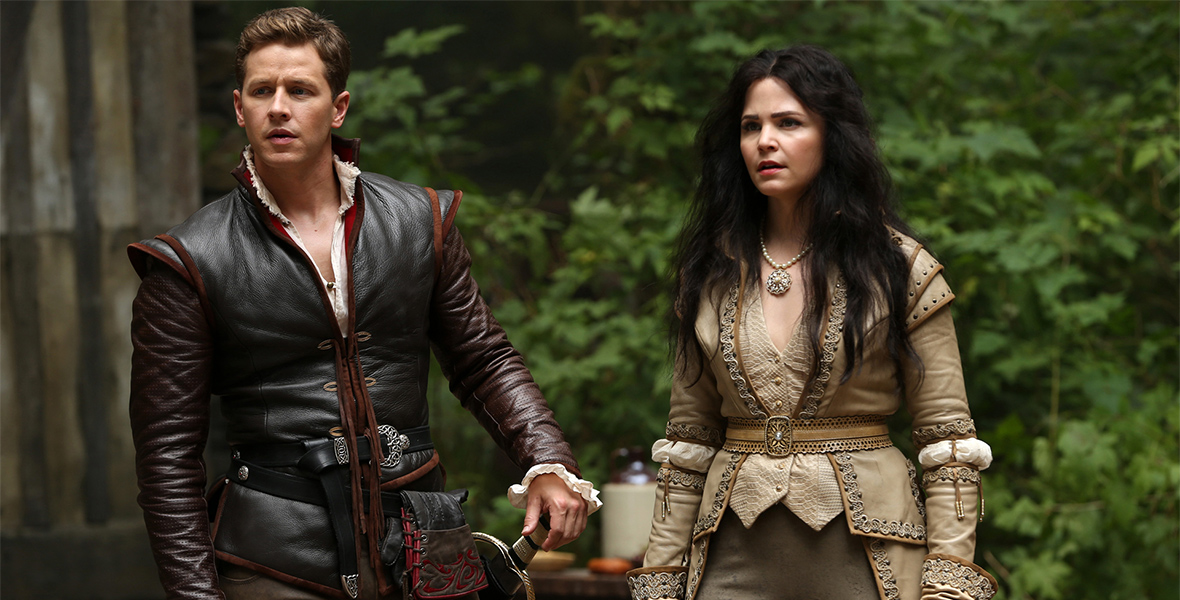 In the forest, Snow White stands beside Prince Charming, looking stoically out at something. Played by Ginnifer Goodwin, Snow White wears an embroidered, belted tan tunic and matching pants. Her black hair is loose and falls past her shoulders. Prince Charming, played by Josh Dallas, wears a long-sleeved, black tunic with red embellishments. His sword sits in its hilt at his side.