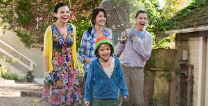 In a still from Ramona and Beezus, Aunt Bea (played by Ginnifer Goodwin) stands outside in a garden with Ramona, Beezus, and their mother. The mother holds an umbrella, though it’s not raining. They’re wet from a water fight, grinning together as they look at something off-camera. Aunt Bea wears a colorful floral dress and a yellow cardigan.