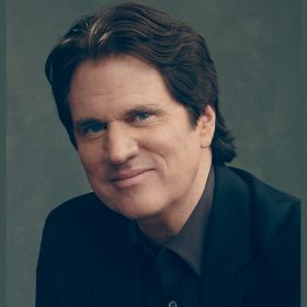 Filmmaker Rob Marshall poses for a photo against a gray-ish green backdrop. He is wearing a black dress shirt and a black blazer and is smiling with his mouth closed.