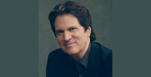 Filmmaker Rob Marshall poses for a photo against a gray-ish green backdrop. He is wearing a black dress shirt and a black blazer and is smiling with his mouth closed.