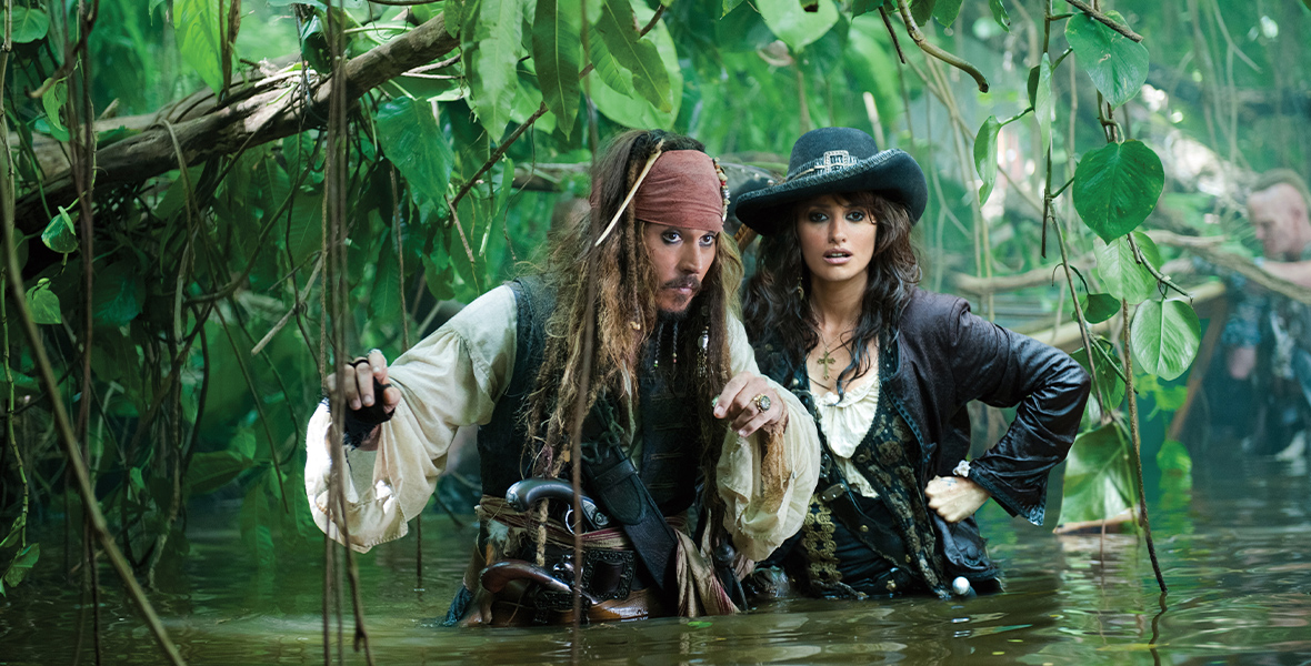 In this production still from Pirates of the Caribbean: On Stranger Tides, Disney Legend Johnny Depp, as Captain Jack Sparrow, and Penelopé Cruz, as Angelica, wade through waist-high water. The two pirates are surrounded by low-hanging trees and vines.