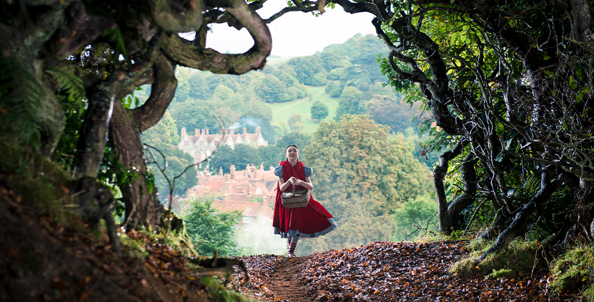 In this production still from Into the Woods, Lilla Crawford, as Little Red Riding Hood, skips along a dirt path while wearing a red cloak and carrying a picnic basket. A quaint valley village is behind her, surrounded by lush trees. In front of her is a canopy.