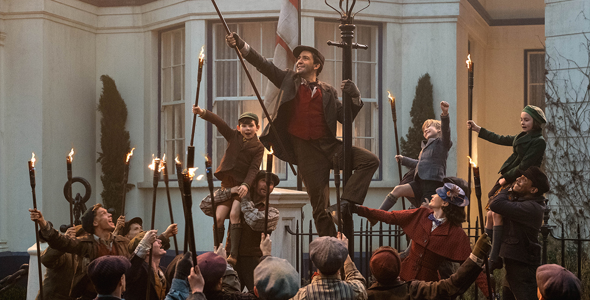 In a production still from Mary Poppins Returns, Lin-Manuel Miranda, as Jack, lifts a torch into the air and wraps an arm and a leg around a lamppost. He is surrounded by three actors portraying the Banks children and a dozen lamplighters, who are holding torches. Emily Blunt, as Mary Poppins, looks up at Jack as she holds onto his lamppost.
