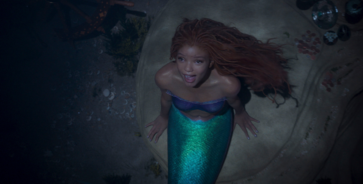 In a production still from The Little Mermaid, Halle Bailey, as Ariel, sits on a rock underwater. She has red dreadlocks, a turquoise tail, and wears a purple bandeau top.