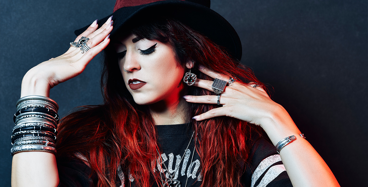 Allison Cimino poses with her eyes closed, one hand behind her ear to show off her earring, while the other rests on the brim of a black hat. She has a variety of jewelery on her fingrs, ears, and neck, and is wearing a black shirt with the word “Disneyland” across it in shiny silver letters.