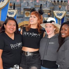 The RockLove team stands in front of Sleeping Beauty Castle at Disneyland. The women on the left of the group photo are all wearing black RockLove tshirts, while the women on the right wear gray RockLove hoodies.