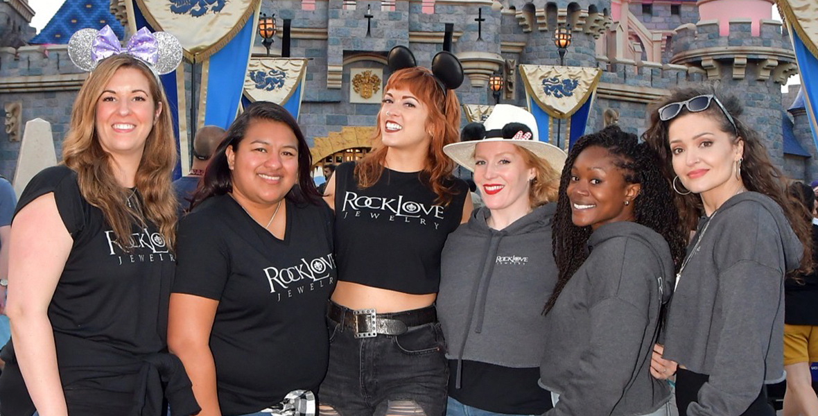 The RockLove team stands in front of Sleeping Beauty Castle at Disneyland. The women on the left of the group photo are all wearing black RockLove tshirts, while the women on the right wear gray RockLove hoodies.