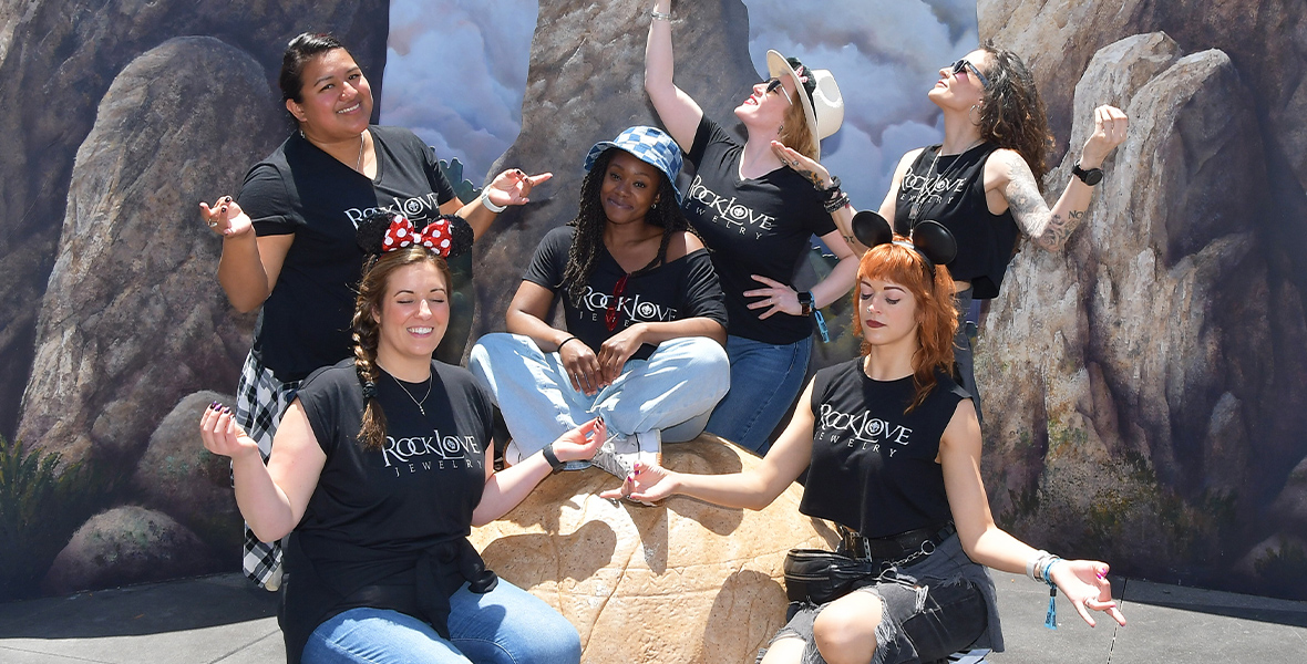 Six members of the RockLove team all strike meditional poses around a big, grey rock. One team member is sitting cross-legged atop the rock while the rest of the women pose around the rock. Everyone is wearing black shirts that have the “RockLove Jewelery” logo in white text across the front.