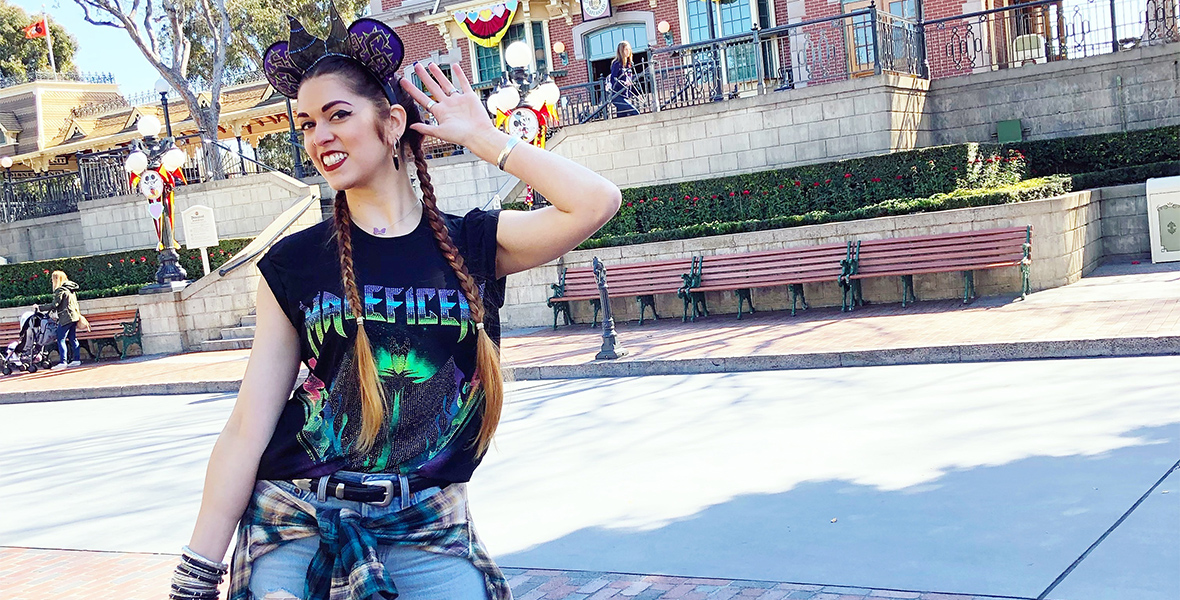 Alison Cimino of RockLove stands in front of the Main Street, U.S.A. train station at Disneyland park. She is wearing an outfit inspired by Maleficent, including a sleeveless t-shirt depicting the character and a coordinating mouse ear headband. She is holding a leather jacket in her right hand and holding her left hand up to her headband.