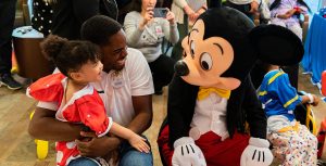 Mickey Mouse sits beside a man and his young daughter, who sits on her father’s lap and wears a Minnie Mouse dress. They both smile at Mickey Mouse.