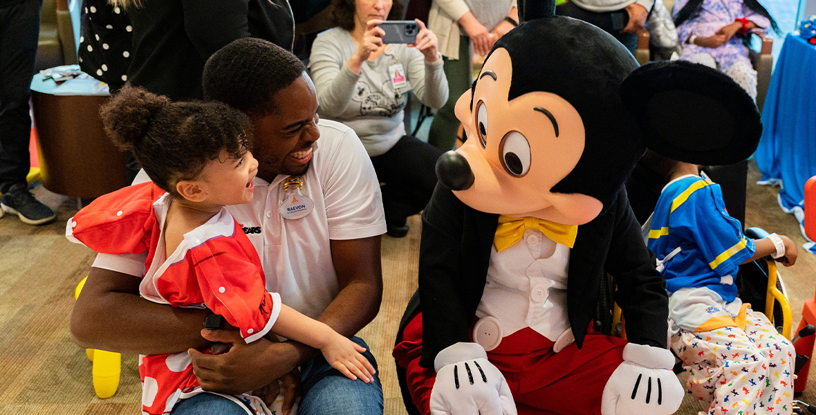 Mickey Mouse sits beside a man and his young daughter, who sits on her father’s lap and wears a Minnie Mouse dress. They both smile at Mickey Mouse.