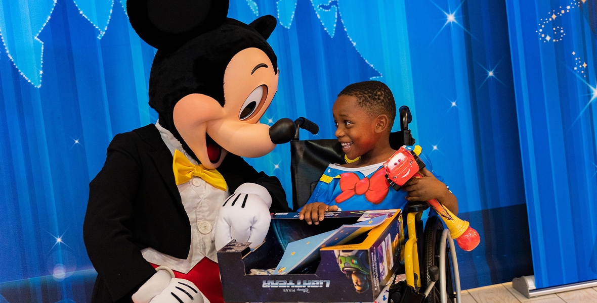 Mickey Mouse kneels beside a child in a wheelchair, who holds a boxed toy and wears Donald Duck hospital wear.