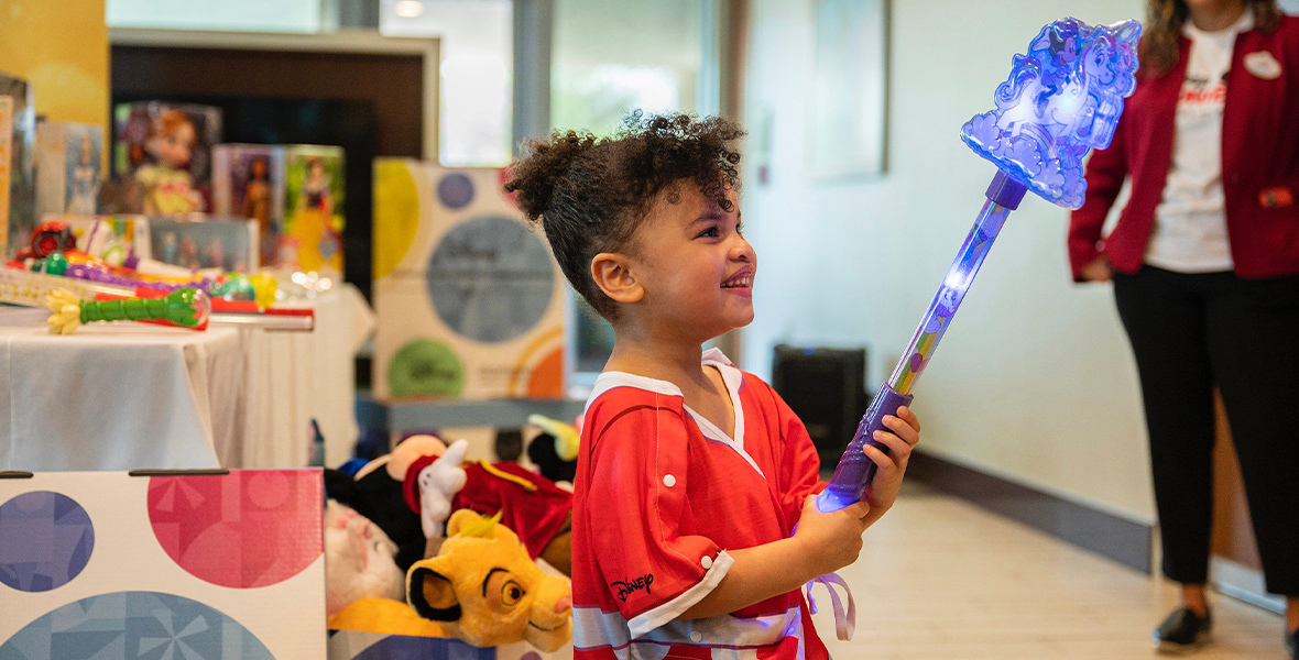 A little girl wearing Minnie Mouse hospital wear waves a blue and purple Frozen wand. In the background, Disney toys like dolls and stuffed animals sit on tables.