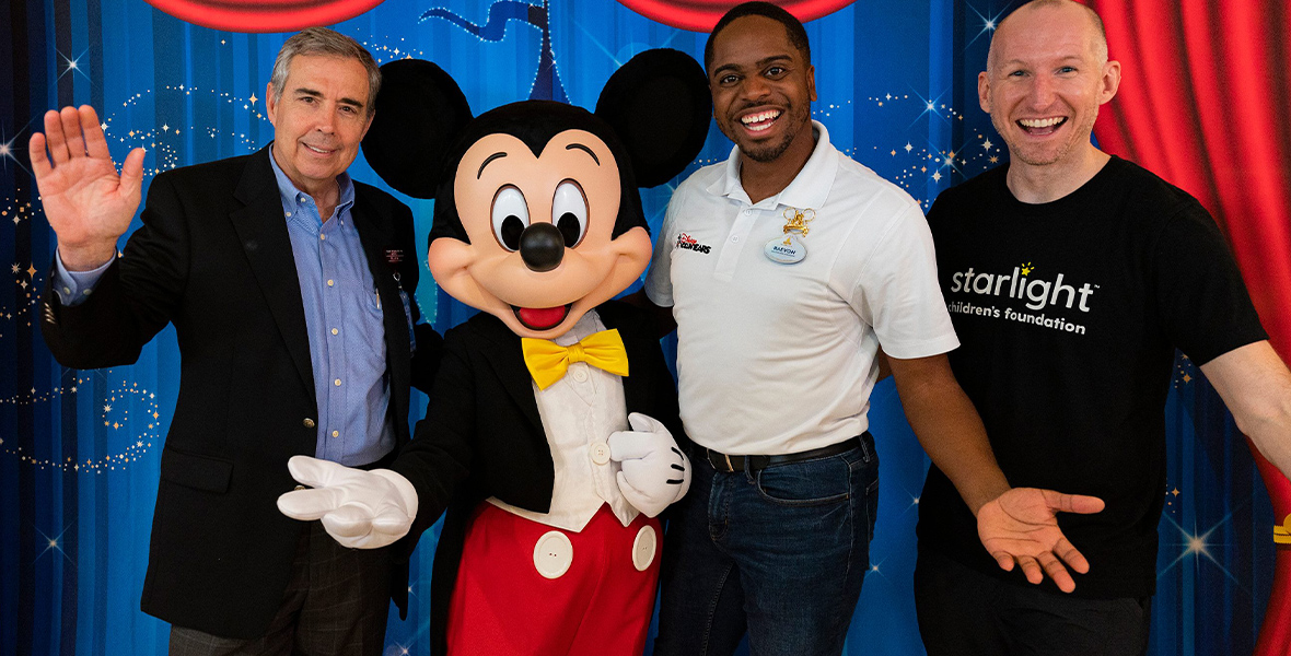 Mickey Mouse poses with three men for the camera. They all smile with their arms in the air while standing in front of a blue curtain.