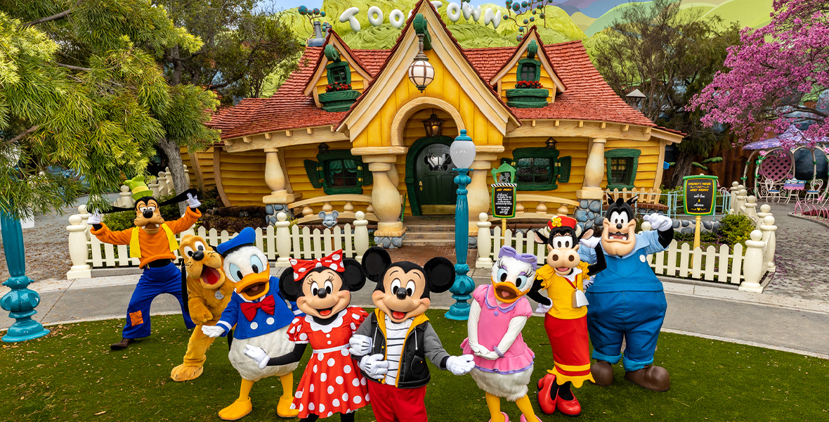 Mickey Mouse, Minnie Mouse, Donald Duck, Daisy Duck, Pluto, Goofy, Clarabelle, and Pete pose together on the lawn outside of Mickey Mouse’s house. The yellow, quirky house is delightfully cartoon-y, while big white letters spell out “Toontown” on the green hills behind it.