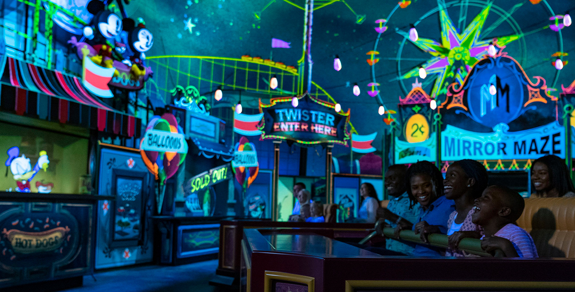 On Mickey & Minnie’s Runaway Railway, guests experience a carnival segment. The stands are lit and whimsical, such as a balloon stand and mirror maze. A bright Ferris wheel glows in the background underneath an indoor starry sky.