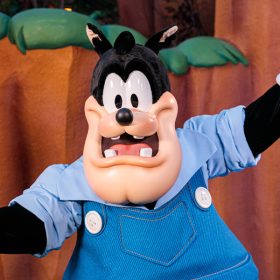 The character Pete stands with his arms out in Mickey's Toontown as he poses for the camera. He wears blue overalls, a blue collared shirt, and white gloves.