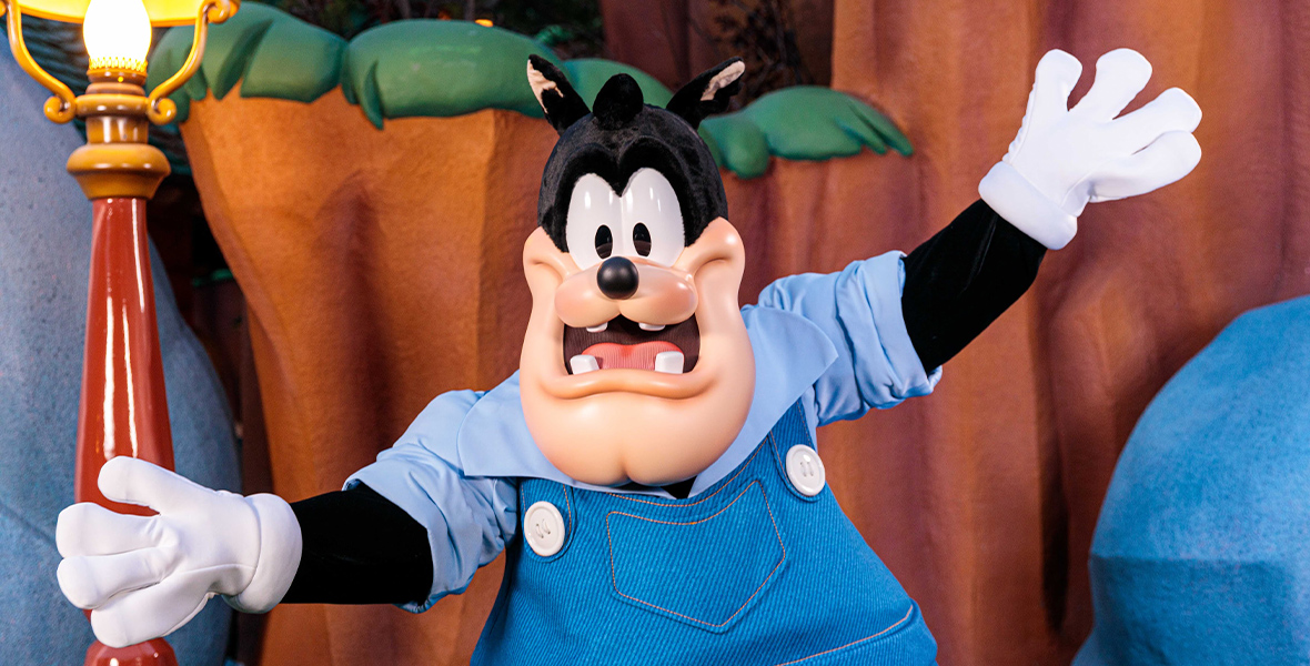 The character Pete stands with his arms out in Mickey's Toontown as he poses for the camera. He wears blue overalls, a blue collared shirt, and white gloves.