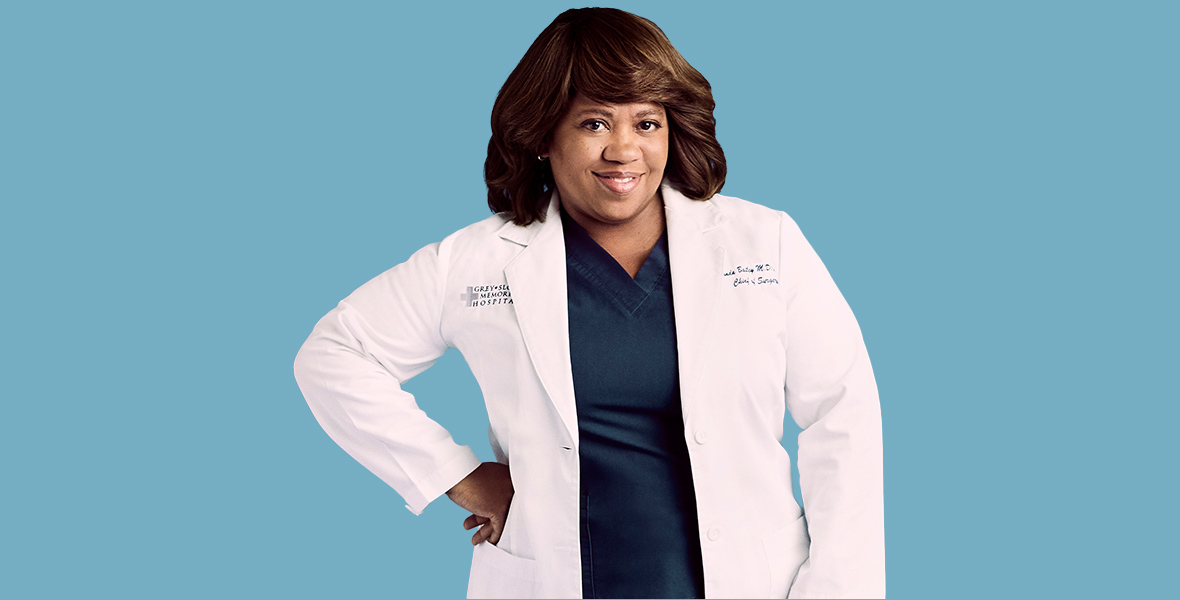 In a promotional photo for Grey’s Anatomy, Chandra Wilson, as Dr. Miranda Bailey, poses against a blue backdrop. She wears navy blue scrubs, black rubber shoes, and a white lab coat that says “Grey + Sloane Memorial Hospital” on the right side of her chest and “Miranda Bailey, M.D., Chief of Surgery” on the left side of her chest.