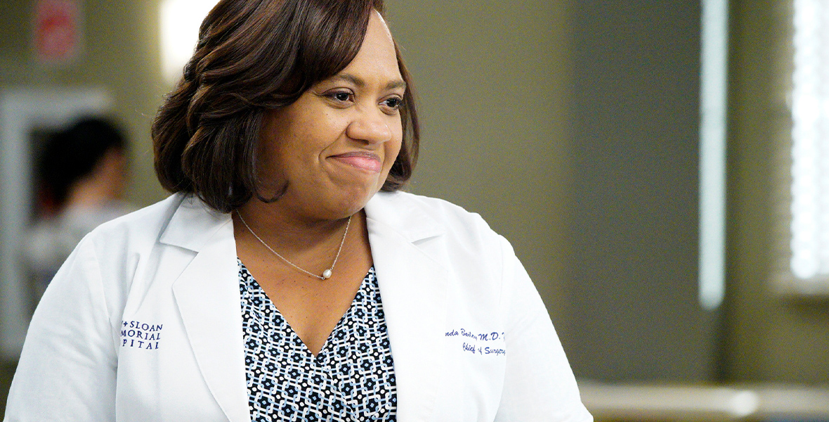 In a production still from Grey’s Anatomy, Chandra Wilson, as Dr. Miranda Bailey, wears a white lab coat that says “Grey + Sloane Memorial Hospital” on the right side of her chest and “Miranda Bailey, M.D., Chief of Surgery” on the left side of her chest. Underneath it, she wears a black, white, and blue wrap top and a jeweled watch.