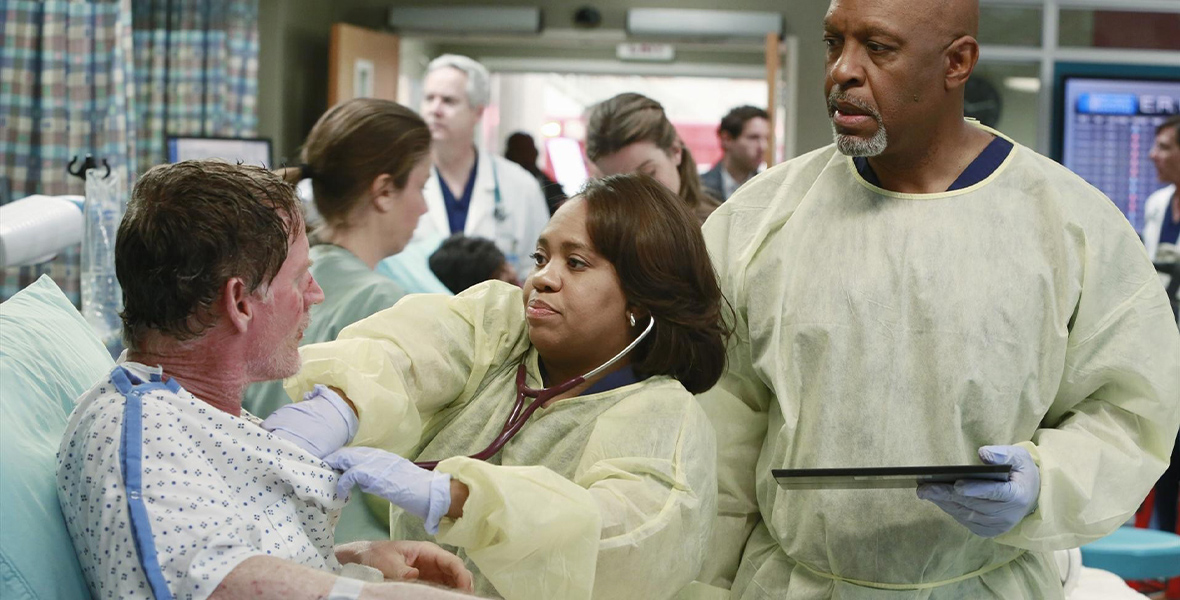 In a production still from Grey’s Anatomy, Chandra Wilson, as Dr. Miranda Bailey, wears a yellow hospital gown over navy blue scrubs. She is using a stethoscope to listen to a patient’s chest. To her left is James Pickens Jr. as Dr. Richard Webber, who is also wearing a yellow hospital gown over navy blue scrubs. He is holding a tablet in his hand. Behind the two doctors are other patients, nurses, and hospital staff.