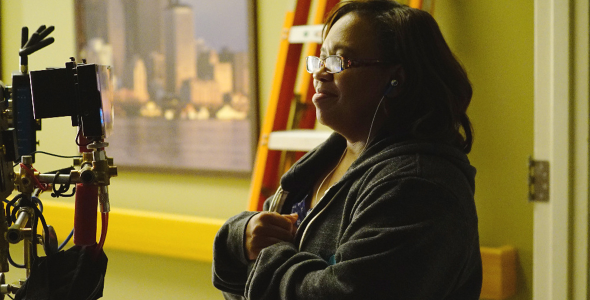 Chandra Wilson watches a monitor while directing a scene from Grey’s Anatomy. She is smiling while wearing headphones, eyeglasses, and a gray hoodie.