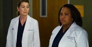 In a production still from Grey’s Anatomy, Disney Legend Ellen Pompeo, as Dr. Meredith Grey, wears navy scrubs under a white lab coat that says “Grey + Sloane Memorial Hospital” on the right side of her chest and “Meredith Grey, M.D.” on the left side of her chest. Her hair is pulled back into a ponytail and she is standing in a hallway. She looks to her left at Chandra Wilson, as Dr. Miranda Bailey, who wears a white lab coat that says “Grey + Sloane Memorial Hospital” on the right side of her chest and “Miranda Bailey, M.D., Chief of Surgery” on the left side of her chest.