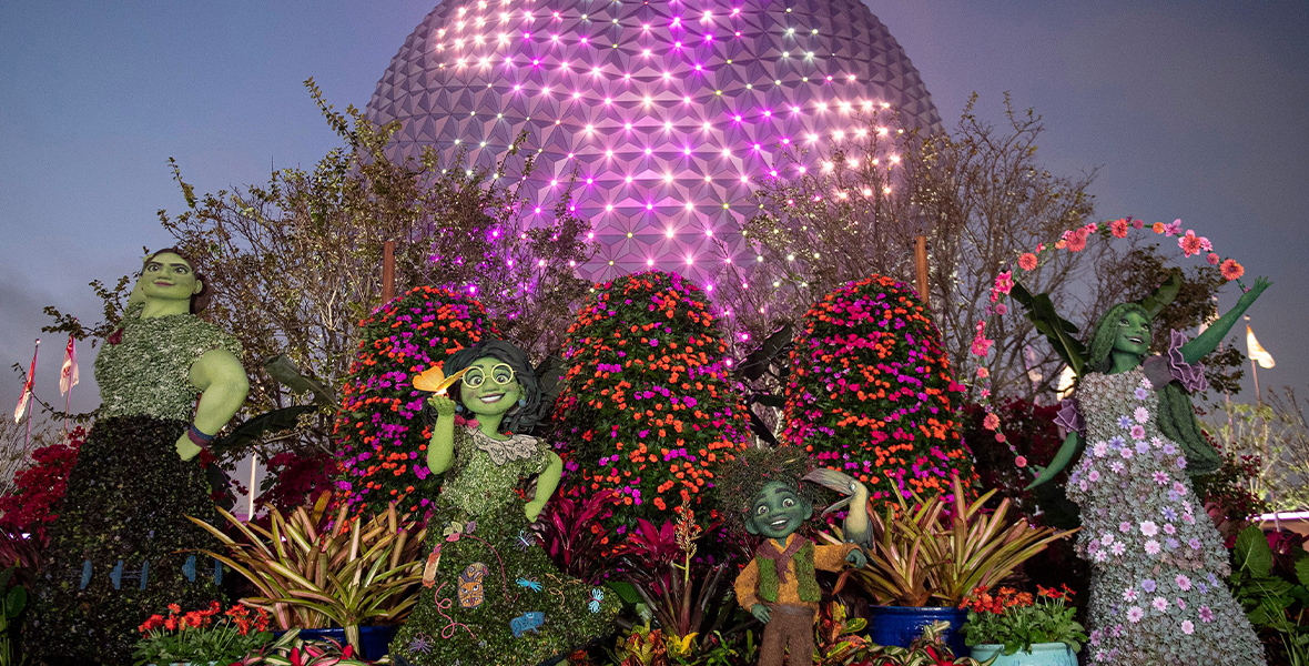 In front of the glowing Spaceship Earth at EPCOT, Encanto topiaries stand amid the greenery. From left to right are topiaries of Luisa, Mirabel, Antonio, and Isabela, clad in festive colors and accessories. Isabela throws up a spray of flowers from her hands, while an orange butterfly rests on Mirabel’s palm.