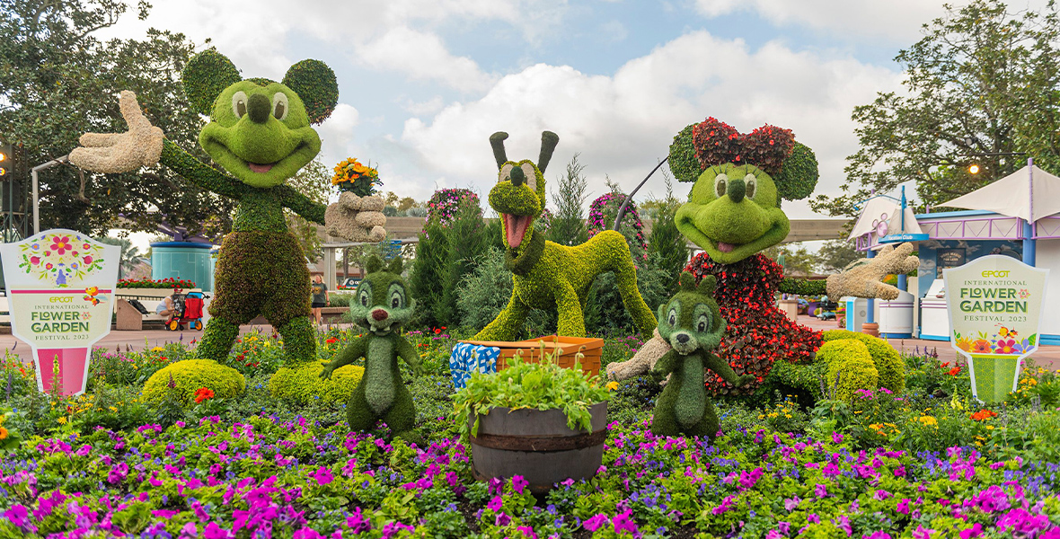 In a sea of flowers, topiaries of Mickey Mouse, Minnie Mouse, Chip, Dale, and Pluto wear welcoming smiles. They’re primarily green, except for Mickey’s white gloves and Minnie’s botanical red dress and bow. Pluto stands on four legs in the center, a picnic basket in front of him.