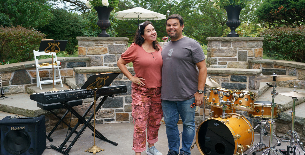 Kristen Anderson-Lopez (left) and Robert Lopez (right) stand together in front of a piano and drums on the set of Up Here. They are dressed casually and smiling.