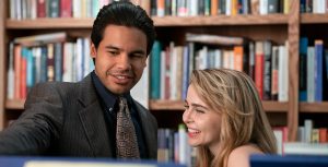In a scene from an episode of Up Here, actors Carlos Valdes and Mae Whitman stand in a bookstore surrounded by colorful books. Valdes wears a brown suit jacket, a blue, button-down shirt, and a tan pattered necktie.