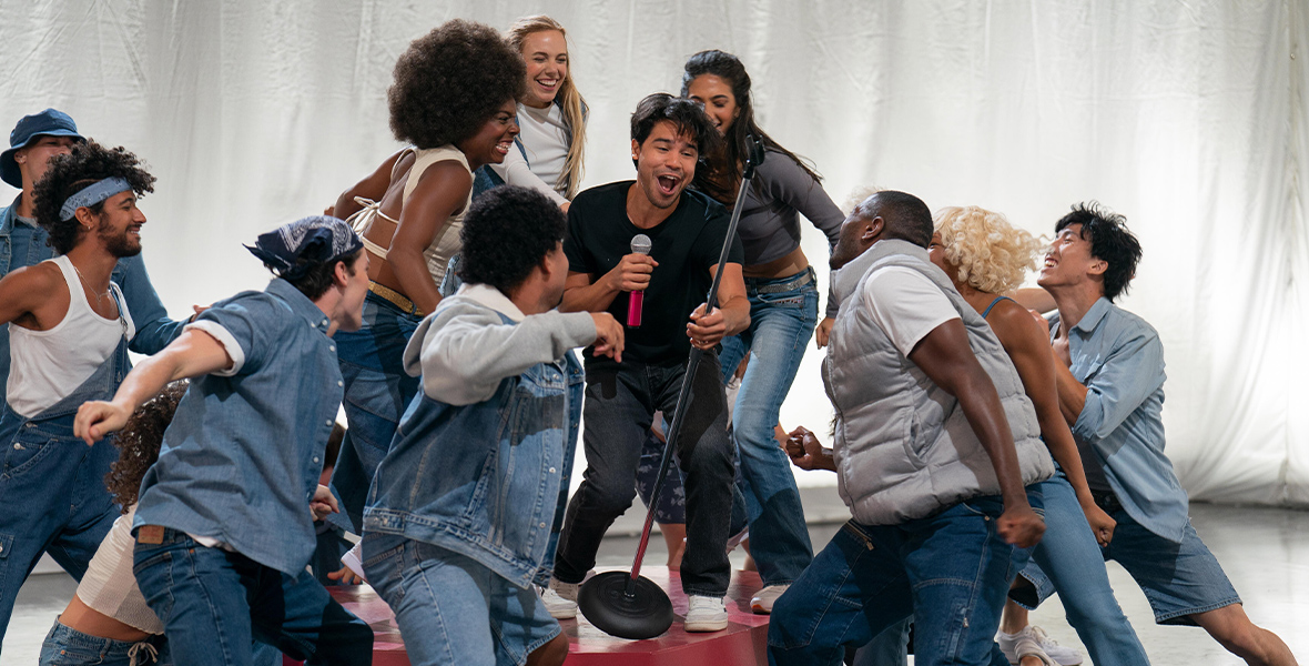 In a scene from an episode of Up Here, actor Carlos Valdes stands on a red platform and holds a microphone stand as dancers surround him. Valdes wears a black T-shirt and black denim jeans.