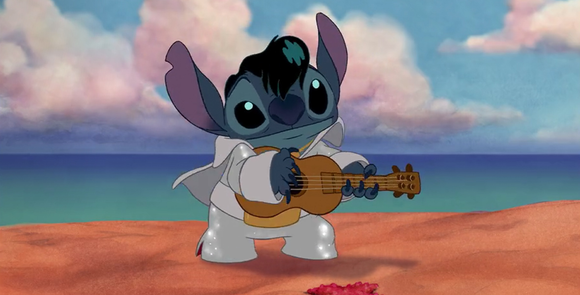 Alt text: In the animated film Lilo and Stitch, the blue alien Stitch stands on the beach in an Elvis Presley costume. He wears a black wig, sparkling white pantsuit, and strums a guitar.